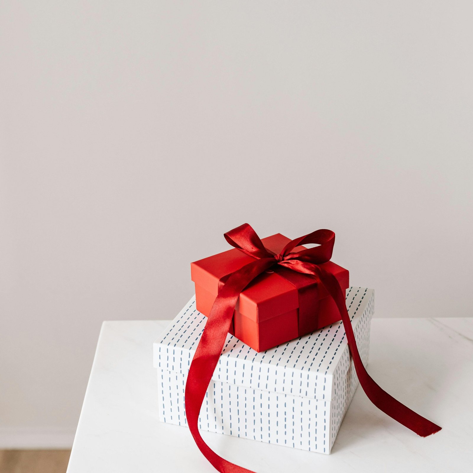 From above of wrapped with ribbon red square gift box placed on white with silver little rhombuses square box on white table against gray background